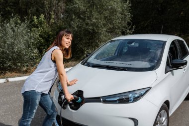 Genç ve güzel bir kadın ormanda bulunan şarj istasyonunda elektrikli arabasını şarj ediyor. Çevre dostu trasport cocnept