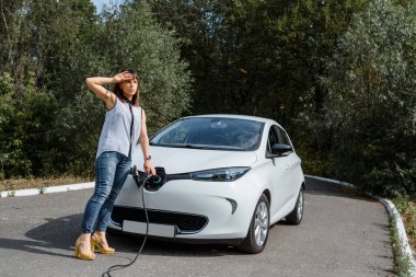 Genç ve güzel bir kadın ormanda bulunan şarj istasyonunda elektrikli arabasını şarj ediyor. Çevre dostu trasport cocnept