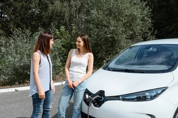 İki mutlu genç kadın, elektrikli arabaları ormandaki şarj istasyonunda şarj olana kadar birbirleriyle konuşuyorlar. Çevre dostu ulaşım konsepti