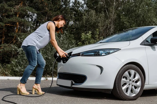 Genç ve güzel bir kadın ormanda bulunan şarj istasyonunda elektrikli arabasını şarj ediyor. Çevre dostu trasport cocnept