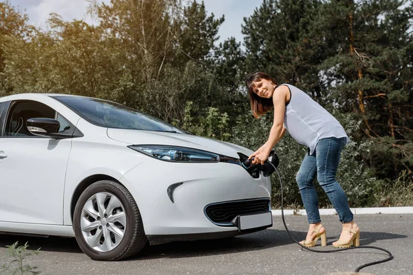 Genç ve güzel bir kadın ormanda bulunan şarj istasyonunda elektrikli arabasını şarj ediyor. Çevre dostu trasport cocnept