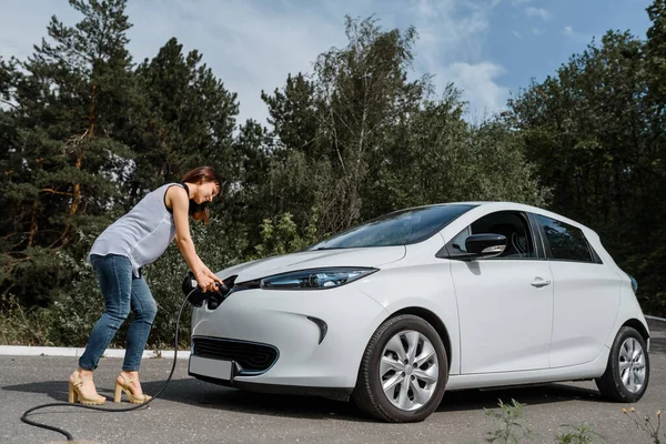 Genç ve güzel bir kadın ormanda bulunan şarj istasyonunda elektrikli arabasını şarj ediyor. Çevre dostu trasport cocnept
