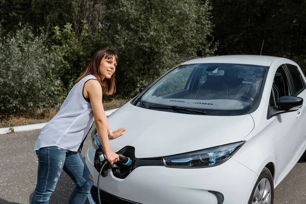 Genç ve güzel bir kadın ormanda bulunan şarj istasyonunda elektrikli arabasını şarj ediyor. Çevre dostu trasport cocnept
