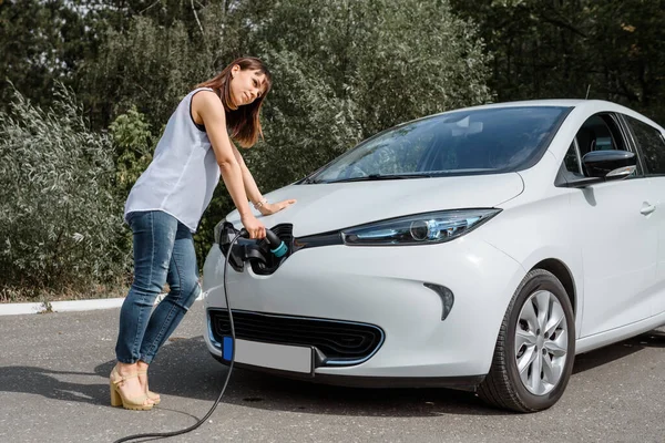 Genç ve güzel bir kadın ormanda bulunan şarj istasyonunda elektrikli arabasını şarj ediyor. Çevre dostu trasport cocnept