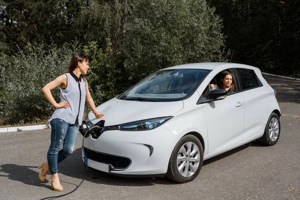 İki mutlu genç kadın, elektrikli arabaları ormandaki şarj istasyonunda şarj olana kadar birbirleriyle konuşuyorlar. Çevre dostu ulaşım kavramı