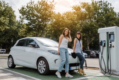 İki mutlu genç kadın, otoparktaki şarj istasyonunda elektrikli arabaları şarj olana kadar birbirleriyle konuşuyorlar. Çevre dostu ulaşım konsepti