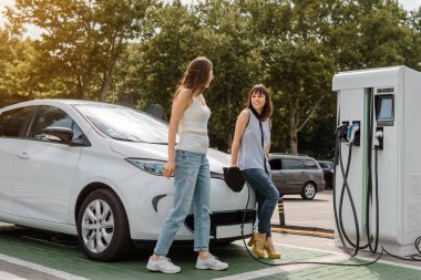 İki mutlu genç kadın, otoparktaki şarj istasyonunda elektrikli arabaları şarj olana kadar birbirleriyle konuşuyorlar. Çevre dostu ulaşım konsepti