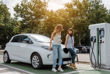 İki mutlu genç kadın, otoparktaki şarj istasyonunda elektrikli arabaları şarj olana kadar birbirleriyle konuşuyorlar. Çevre dostu ulaşım konsepti