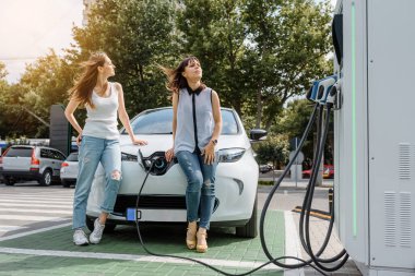 İki mutlu genç kadın, otoparktaki şarj istasyonunda elektrikli arabaları şarj olana kadar birbirleriyle konuşuyorlar. Çevre dostu ulaşım konsepti