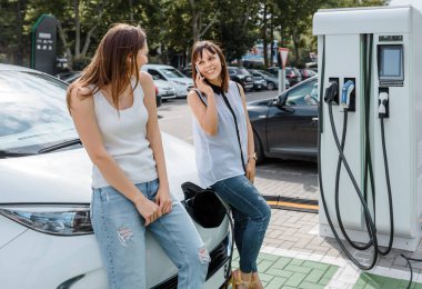 İki mutlu genç kadın, otoparktaki şarj istasyonunda elektrikli arabaları şarj olana kadar birbirleriyle konuşuyorlar. Çevre dostu ulaşım kavramı