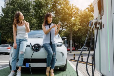 İki kadının en iyi arkadaşı elektrikli arabasının parasını ödemek için akıllı telefonunu kullanana kadar elektrikli arabada oturuyorlar. Mobil ödeme konsepti. Çevre dostu elektrikli araba teknolojisi