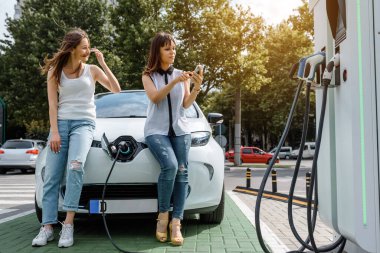 İki kadının en iyi arkadaşı elektrikli arabasının parasını ödemek için akıllı telefonunu kullanana kadar elektrikli arabada oturuyorlar. Mobil ödeme konsepti. Çevre dostu elektrikli araba teknolojisi