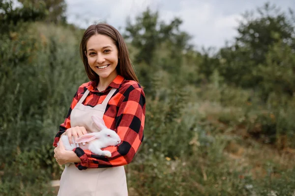 Młoda Uśmiechnięta Farmerka Patrzy Obiektyw Trzyma Białego Królika Zewnątrz Bio — Zdjęcie stockowe