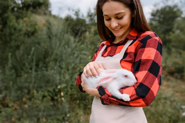 Młoda Uśmiechnięta Farmerka Trzyma Rękach Białego Królika Patrzy Niego Zewnątrz — Zdjęcie stockowe