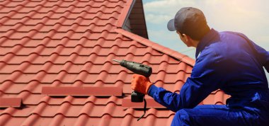 Back view of contractor worker in blue overalls is repairing a red roof with electric screw driver.  Roofing concept. Copy space for you text clipart