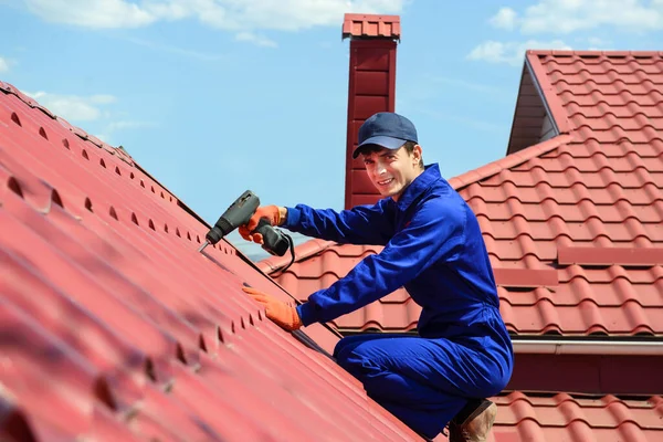 Young happy man contractor worker in blue overalls is repairing a red roof with electric screw driver. He is smiling and looking to the camera. Roofing concept. Copy space for you text