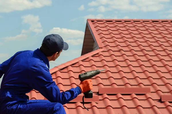 Back view of contractor worker in blue overalls is repairing a red roof with electric screw driver.  Roofing concept. Copy space for you text