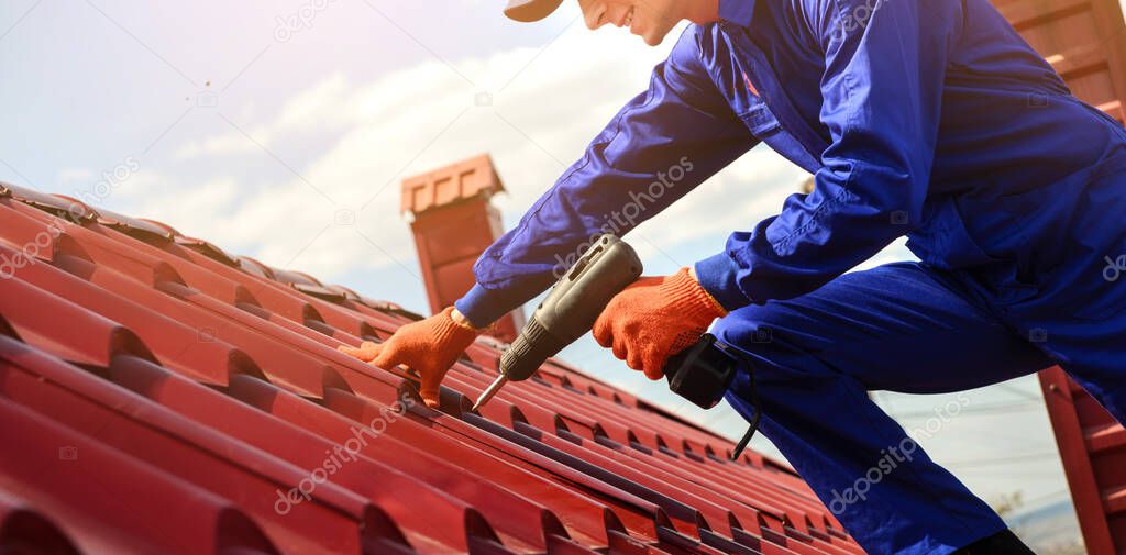 Close up of young happy man contractor worker in blue overalls is repairing a red roof with electric screw driver. He is smiling. Roofing concept. Copy space for you text