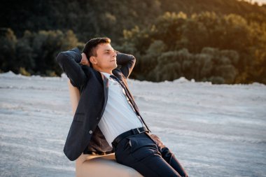 Young man in a classic jacket is sitting on the chair with hands behind his head and making a rest. Concept stop working, you need a vacation.