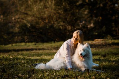 Young beautiful woman in white shirt is hugging her white dog samoyed outdoors in the park and sitting on the grass.