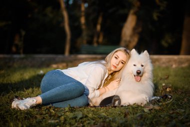 Young beautiful woman in white shirt is hugging her white dog samoyed outdoors in the park and sitting on the grass.