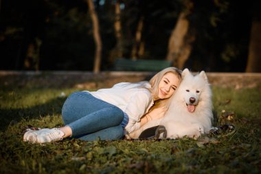 Young beautiful woman in white shirt is hugging her white dog samoyed outdoors in the park and sitting on the grass.