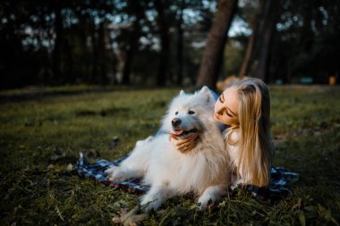 Young beautiful woman in white shirt is hugging her white dog samoyed outdoors in the park and sitting on the grass.