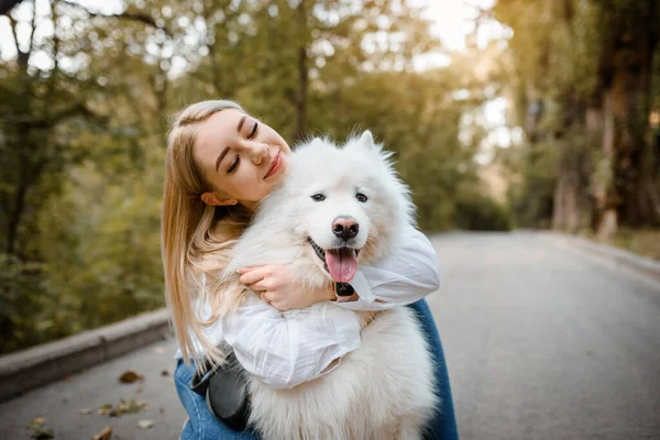 Beyaz gömlekli genç bir kadın parkta beyaz köpeğini kucaklıyor..
