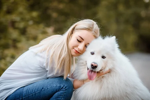 穿着白衬衫的年轻漂亮的女人正在公园里抱着自己的白狗 — 图库照片