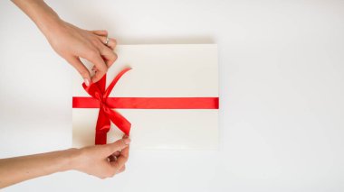 Female hands holding a gift box white with a red ribbon, one object on a white background. Gift holiday. Festive background. Top View. Free space for text