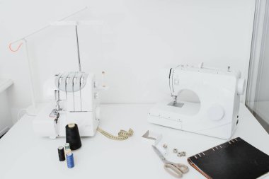 Creativ tailor workplace. Close up sewing and overlock machines, spool of thread, scissors, centimeter ribbon and black notebook on the table