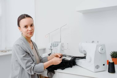 Young woman tailor is sewing piece of black lace fabric in her white mini workshop and preparing to create sexy underwear. On the table are sewing and overlock machines with spools of thread.