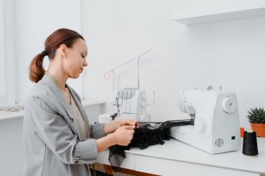 Young woman tailor is sewing piece of black lace fabric in her white mini workshop and preparing to create sexy underwear. On the table are sewing and overlock machines with spools of thread.
