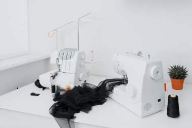 Creativ tailor workplace. Close up sewing and overlock machines, spool of thread, scissors, centimeter ribbon and pieces of black material on the table