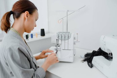 Tailor work space concept. Female tailor with a centimeter ribbon over the neck is adjusting the overlock machine/ serger on the table in her mini tailoring workshop. On the table are sewing machin
