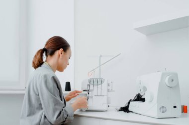 Tailor work space concept. Female tailor with a centimeter ribbon over the neck is adjusting the overlock machine/ serger on the table in her mini tailoring workshop. On the table are sewing machin