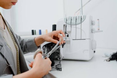 Young woman tailor is cutting with scissors a piece of black material in her white mini workshop. On the table are sewing and overlock machines with spools of thread.