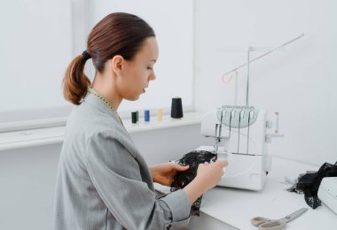 Young woman tailor is looking at a piece of black material in her white mini workshop and preparing to create underwear. On the table are sewing and overlock machines with spools of thread.
