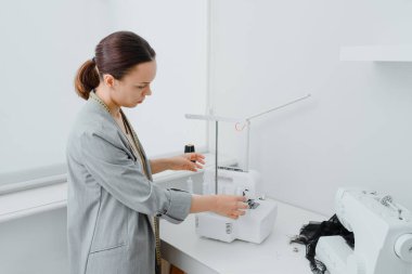 Young woman tailor with centimeter ribbon over the neck is adjusting the overlock machine/ serger on the table in her mini tailoring workshop