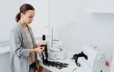 Young woman tailor is looking at a piece of black material in her white mini workshop and preparing to create underwear. On the table are sewing and overlock machines with spools of thread.