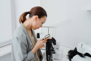 Young woman tailor is looking at a piece of black material through a magnifying glass in her white mini workshop and preparing to create underwear. On the table are sewing and overlock machines with spools of thread.