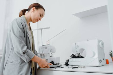 Young woman tailor is cutting with scissors a piece of black material in her white mini workshop. On the table are sewing and overlock machines with spools of thread.