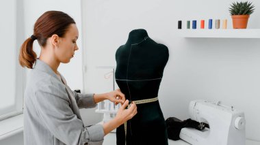 Young woman tailor is measuring with a centimeter ribbon the waist of a mannequin in her mini workshop for creating new clothes.