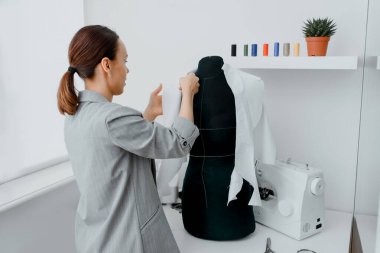Young woman tailor is arranging a future shirt on a mannequin in her mini workshop. On the table are: overlock  and sewing machine. Creative design concept.