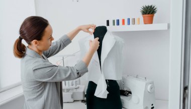 Young woman tailor is arranging a future shirt on a mannequin in her mini workshop. On the table are: overlock  and sewing machine. Creative design concept.