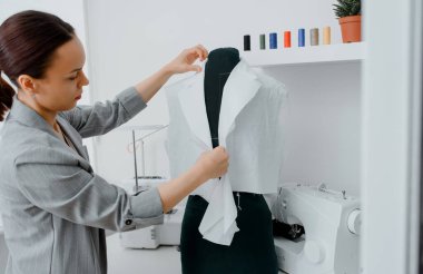 Young woman tailor is arranging a future shirt on a mannequin in her mini workshop. On the table are: overlock  and sewing machine. Creative design concept.