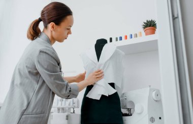 Young woman tailor is arranging a future shirt on a mannequin in her mini workshop. On the table are: overlock  and sewing machine. Creative design concept.