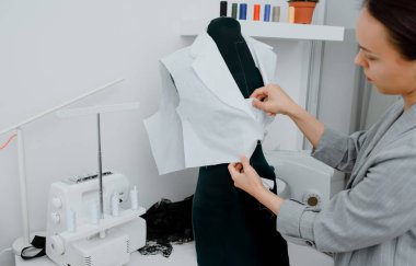 Young woman tailor is arranging a future shirt on a mannequin in her mini workshop. On the table are: overlock  and sewing machine. Creative design concept.