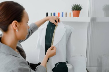 Young woman tailor is measuring with a centimeter ribbon a future shirt on a mannequin in her mini workshop