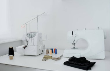 Creativ tailor workplace. Close up sewing and overlock machines, spool of thread, scissors, centimeter ribbon and pieces of black material on the table